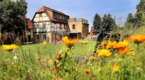 A traditional half-timbered building with a sloped roof, surrounded by a grassy field with wildflowers. - Exclusive 4-star holiday home for 8-12 people on historic Dreiseithof (Pirna)