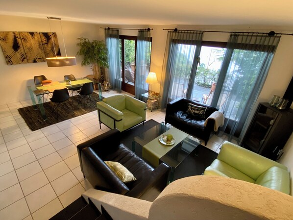 Un salon moderne avec une table basse en verre, un canapé, un coin repas avec une table et des chaises, et une grande fenêtre avec des rideaux.