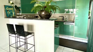 Una cocina con armarios verdes, una isla central con dos taburetes negros, una planta en maceta y un cartel en la pared.