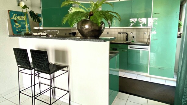 A kitchen with green cabinets, a central island with two black bar stools, a potted plant, and a sign on the wall.