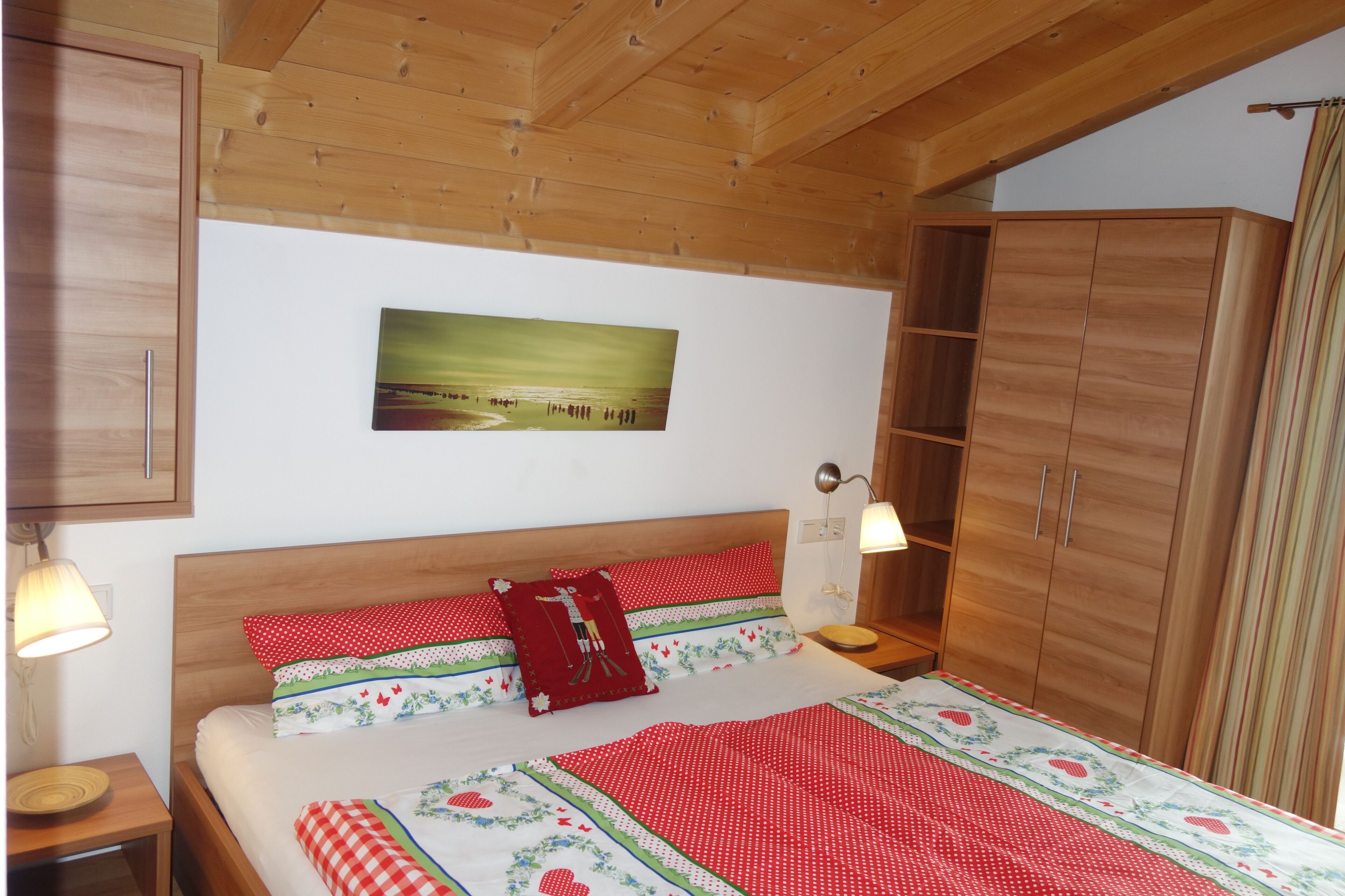 A bedroom with a wooden ceiling, a bed with red and white bedding, a wooden wardrobe, and a nightstand with a lamp.