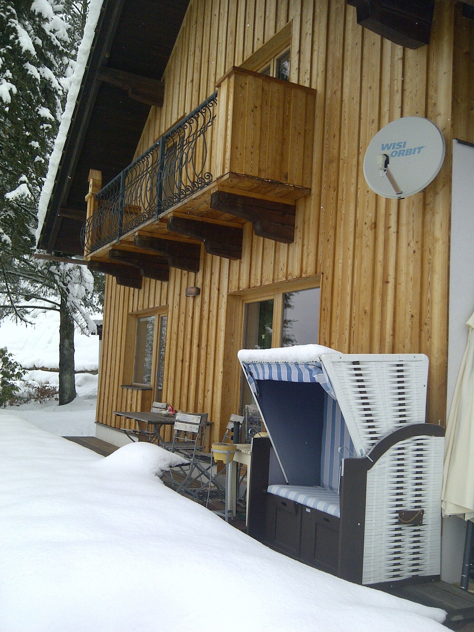 A wooden cabin with a balcony, a satellite dish, and outdoor furniture covered in snow.