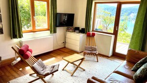 Una sala de estar acogedora con piso de madera, un sofá de cuero marrón, una silla de madera con cojín, una mesita y un televisor en la pared.
