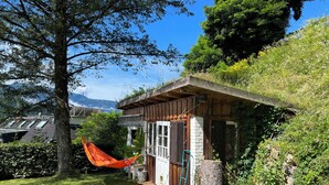 Una pequeña cabaña de madera con techo inclinado, rodeada de exuberante vegetación y una hamaca.