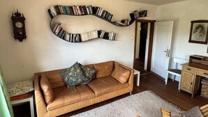 Una sala de estar acogedora con un sofá de cuero marrón, una estantería llena de libros, un mueble de madera y un reloj en la pared.