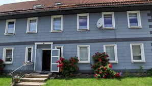 Un edificio de dos pisos con fachada gris, múltiples ventanas con marcos blancos, una antena parabólica y un pequeño jardín con flores rojas.