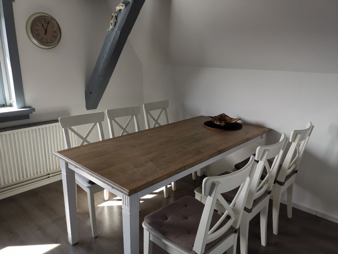 Een eetgedeelte met een houten tafel en witte stoelen, een klok aan de muur en een raam met zonwering.