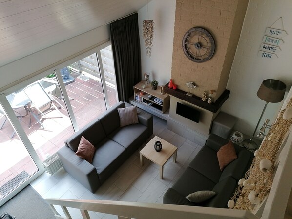 A modern living room with a grey sofa, a coffee table, and a TV mounted on a wall. There is a large window providing a view of the outside.
