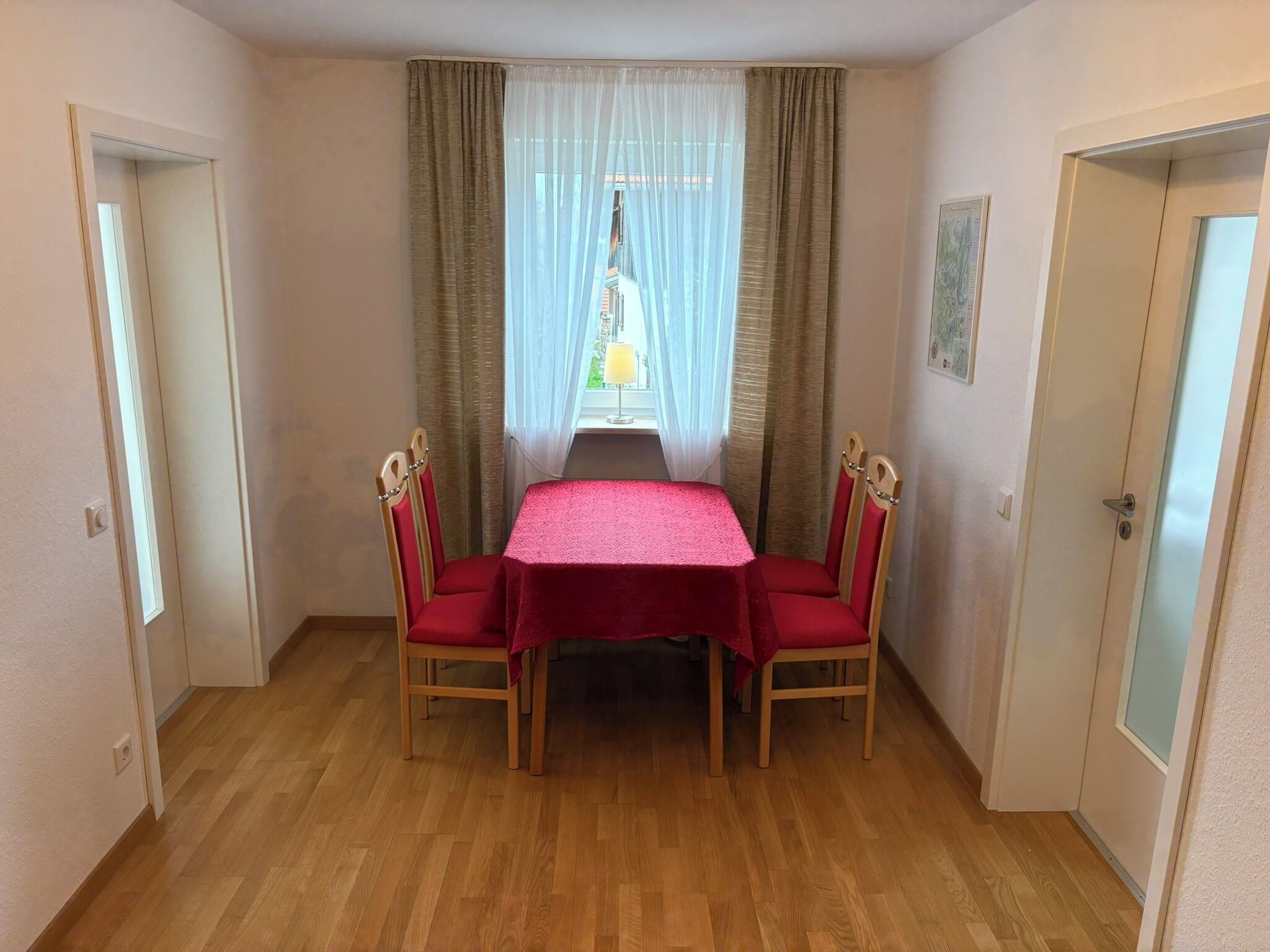 A room with a wooden floor, a table with a red tablecloth, two chairs, and a window with curtains.