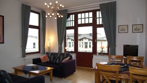 A living room with a sofa, a dining table with chairs, a chandelier, and large windows with curtains. - Villa Medici Apartment 5 (Heringsdorf)