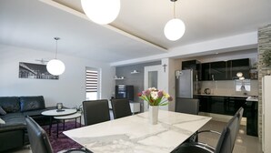 En moderne stue med spisebord, sofa og lænestole, et køkken med sorte skabe, og udsigt over bybilledet.