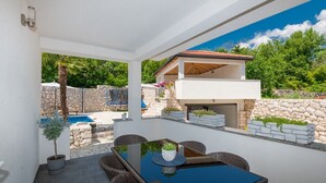 En moderne udendørs terrasse med et glasbord og fletstole, en stenvæg og en pool.