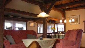 Una sala de estar acogedora con una mesa de madera, dos sofás y un sillón. Hay ventanas con cortinas, una araña y un cuadro en la pared.