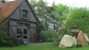 Una casa de madera rústica con techo inclinado, rodeada de abundante vegetación y grandes rocas.