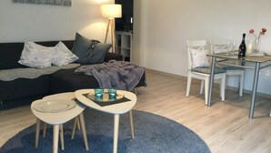 Una sala de estar con un sofá, una mesa de centro y una mesa de comedor con sillas. Hay una lámpara, un cuadro en la pared y una alfombra azul.