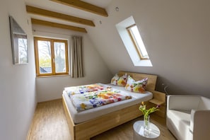 Une chambre à coucher avec un lit en bois, une lucarne, une chaise blanche et une petite table sur laquelle est posé un vase.