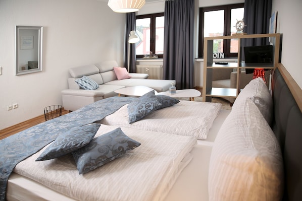Quarto de hotel moderno com uma cama, um sofá, uma mesinha e uma TV sobre um suporte de madeira.