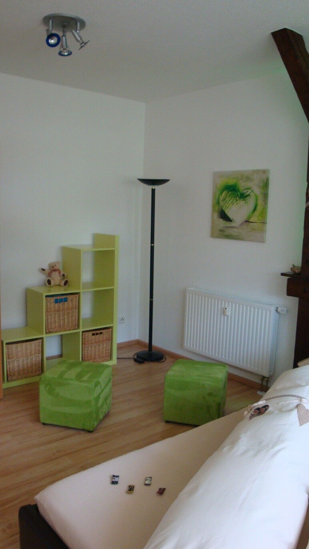 Quarto com cama, pufes verdes, luminária de chão, radiador e prateleiras com cestos.