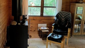 A cozy cabin interior with a wood stove, a chair, a small table, and a window with curtains.