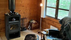 A cozy cabin with a wood-burning stove, a window with a view of greenery, a leather sofa, and a small table.