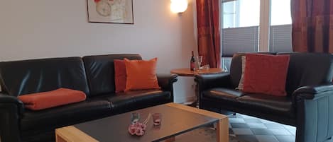 Een woonkamer met twee zwarte leren banken, een houten salontafel, een klein rond tafeltje met een fles en glas, en een kunstwerk in een lijst aan de muur.