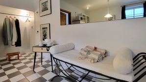 Una habitación con piso a cuadros, una cama con colchón y almohadas blancas, una mesita de noche con lámpara, un espejo y un estante con ropa.