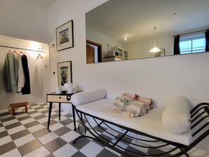 A room with a checkered floor, a bed with a white mattress and pillows, a bedside table with a lamp, a mirror, and a wall-mounted shelf with clothes. - Haus zum Strand 2. 4. - House to the beach 2.4. (Wustrow)