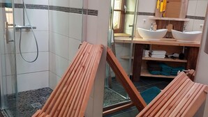 A bathroom with a glass shower enclosure, wooden lounge chairs, a sink, and a mirror.