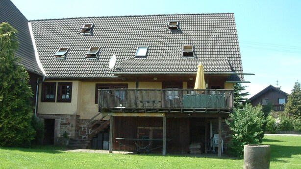 A two-story building with a tiled roof, a balcony with a yellow umbrella, and a stone foundation. - Former farmhouse in a quiet outskirts. Pets are welcome. (Aichhalden)