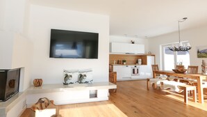 A modern living room with a flat-screen TV mounted on the wall, a wooden dining table with chairs, and a kitchen area in the background. - New holiday home in the mountains, Bad Mitterndorf, Sonnenalm (Bad Mitterndorf)