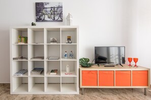 Un salon avec une bibliothèque blanche, un téléviseur posé sur un meuble en bois et un tableau représentant un vélo accroché au mur.