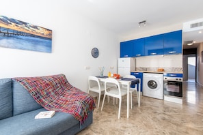 Une cuisine moderne avec des meubles bleus, un coin repas avec des chaises blanches, un canapé recouvert d’un plaid coloré et un tableau représentant un coucher de soleil sur l’eau.