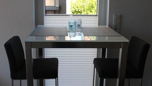 A dining area with a table and two chairs, a window with blinds, and a view of a residential area. - De Hunk, the small apartment in the town center (Borkum)