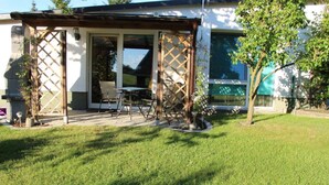 En overdækket terrasse med udendørs møbler, en træpergola og udsigt til et hus med et stort vindue.