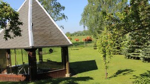 En pavillon med et sekskantet tag og glasvægge, omgivet af græs og træer.