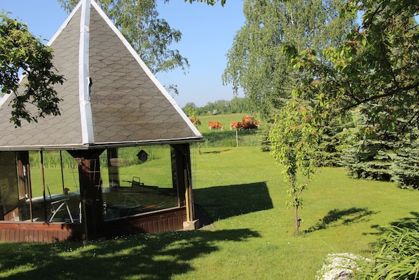 A gazebo with a hexagonal roof and glass walls, surrounded by a grassy area and trees.