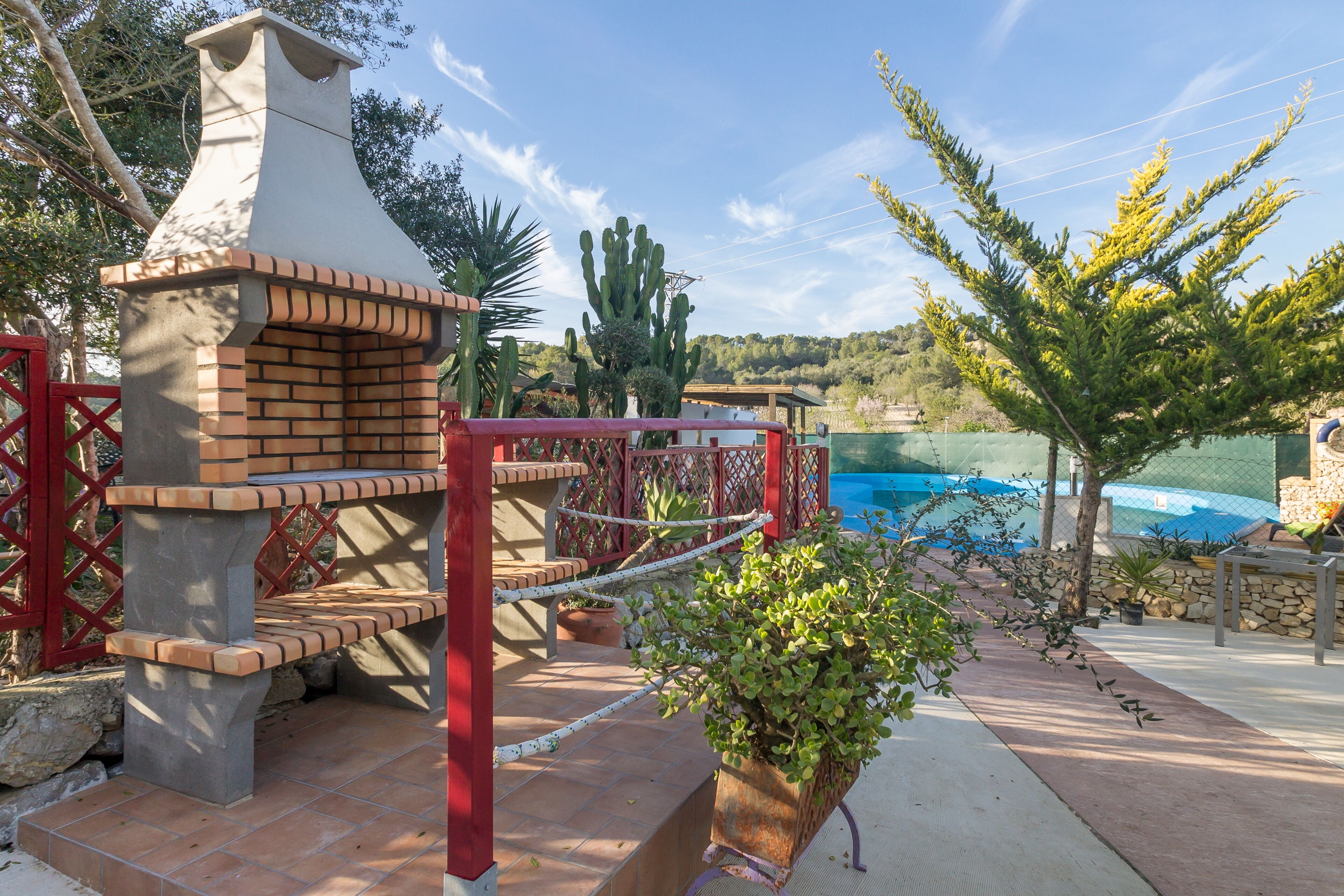 Cucina esterna in mattoni con camino, cancello rosso e piscina sullo sfondo.