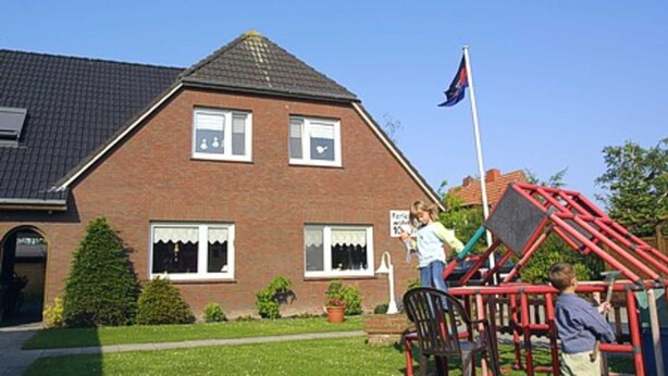 Une maison à deux étages, construite en briques, avec un toit en pente, un mât sur lequel flotte un drapeau, et une aire de jeux avec des équipements rouges.