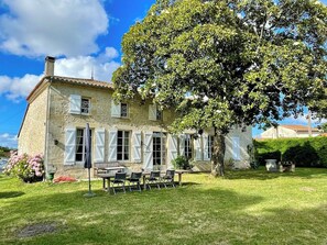 A stone house with a large tree in the yard, a table with chairs, and a small garden.