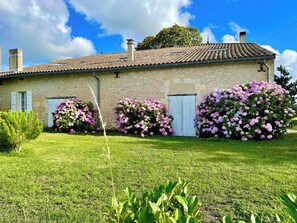 Un bâtiment en pierre avec un toit en tuiles, entouré d’une pelouse bien entretenue et d’arbustes en fleurs.