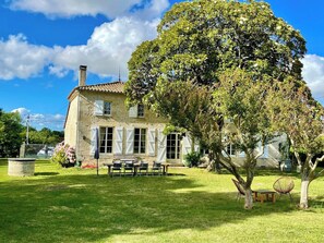 Une maison en pierre avec un toit de tuiles, une cour gazonnée et un grand arbre.