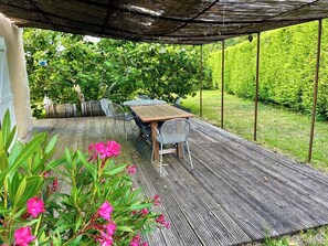 Un patio al aire libre con mesa y sillas de madera, rodeado de follaje y una zona cubierta.