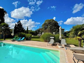 Une piscine entourée de verdure, avec des chaises longues et des parasols, sous un ciel dégagé.