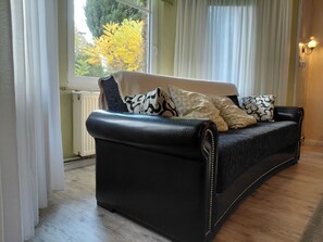 En sort lædersofa med mønstrede puder, et vindue med lette gardiner og en radiator.
