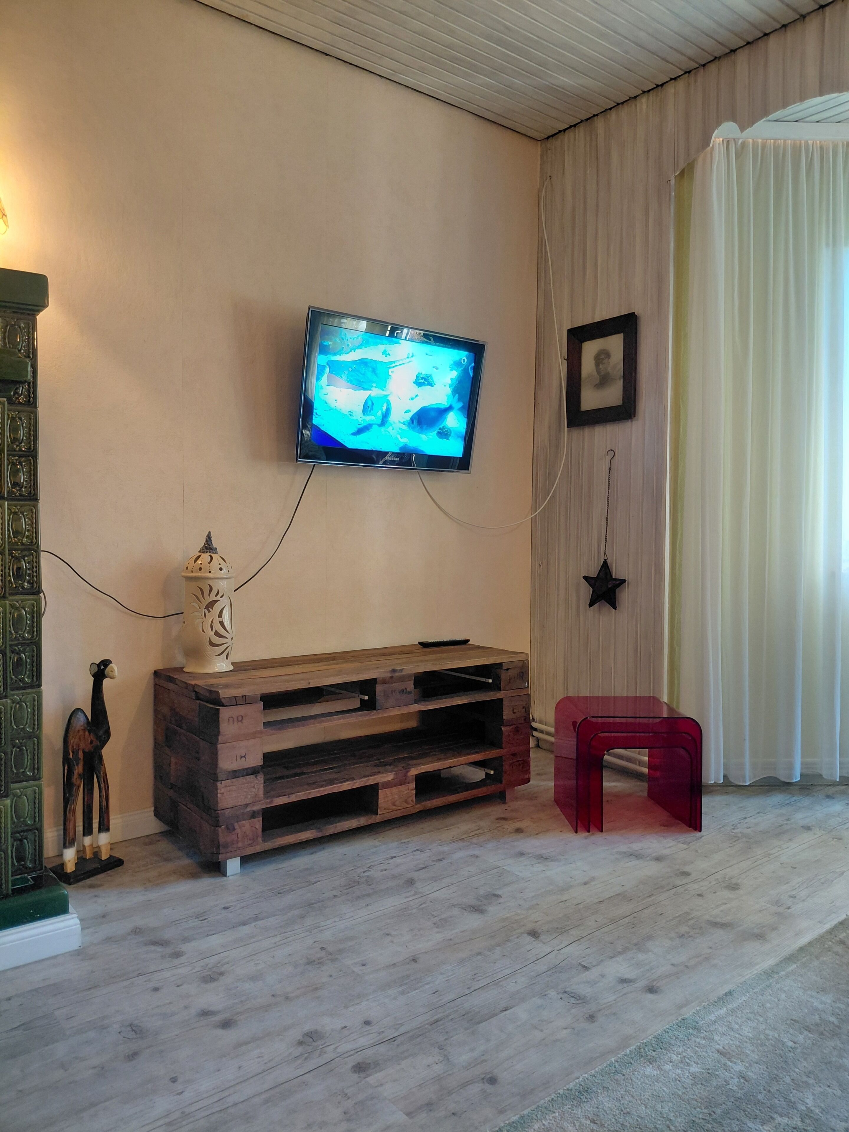 A room with a television mounted on the wall, a wooden entertainment unit, a red stool, and a decorative giraffe figurine.