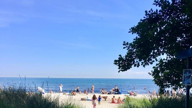 En sti langs stranden fører til en sandstrand med mennesker og parasoller.