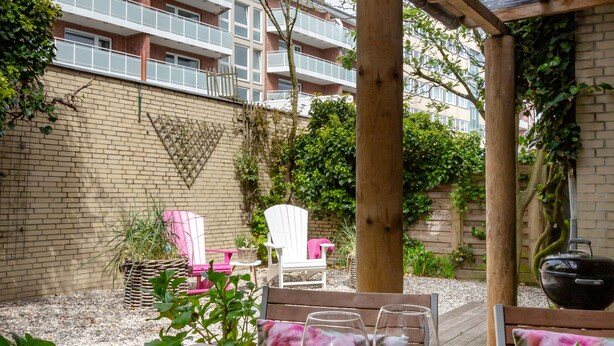 An outdoor patio with a wooden table set for dining, surrounded by a brick wall and a covered area with a view of a residential building. - "Sea lights" apartment on Sylt for 2 people with garden in the city center (Sylt OT Westerland)