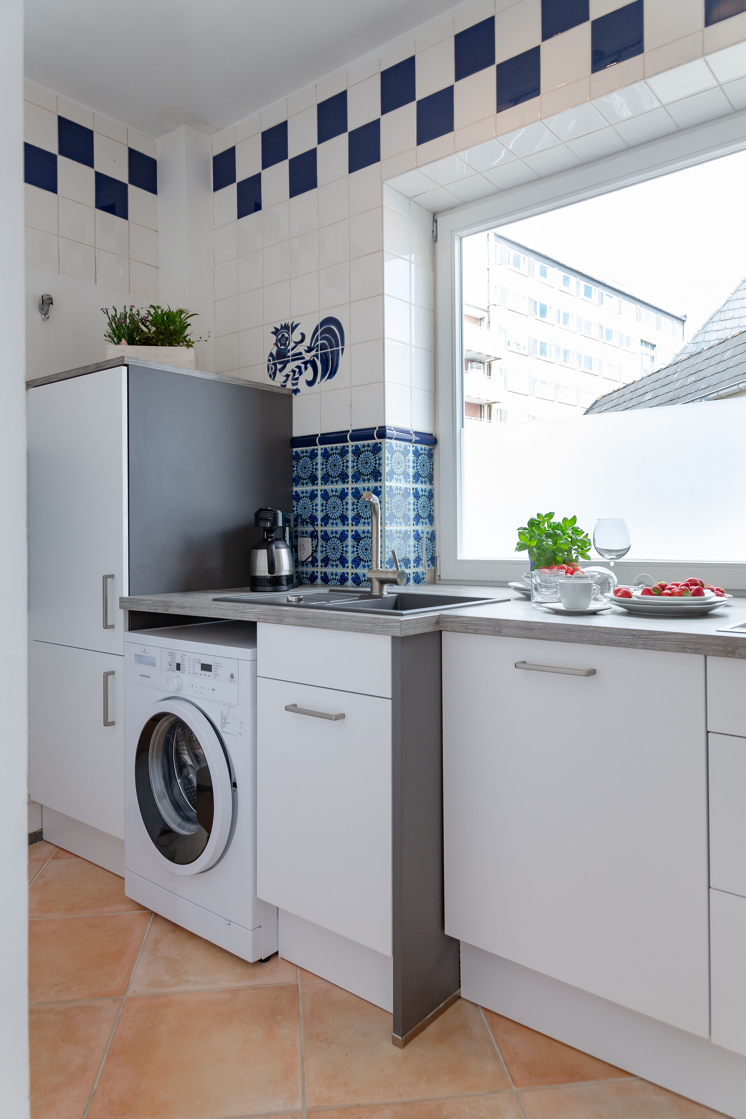 Une cuisine moderne avec des meubles blancs, un lave-linge, une machine à café et une fenêtre donnant sur des immeubles.