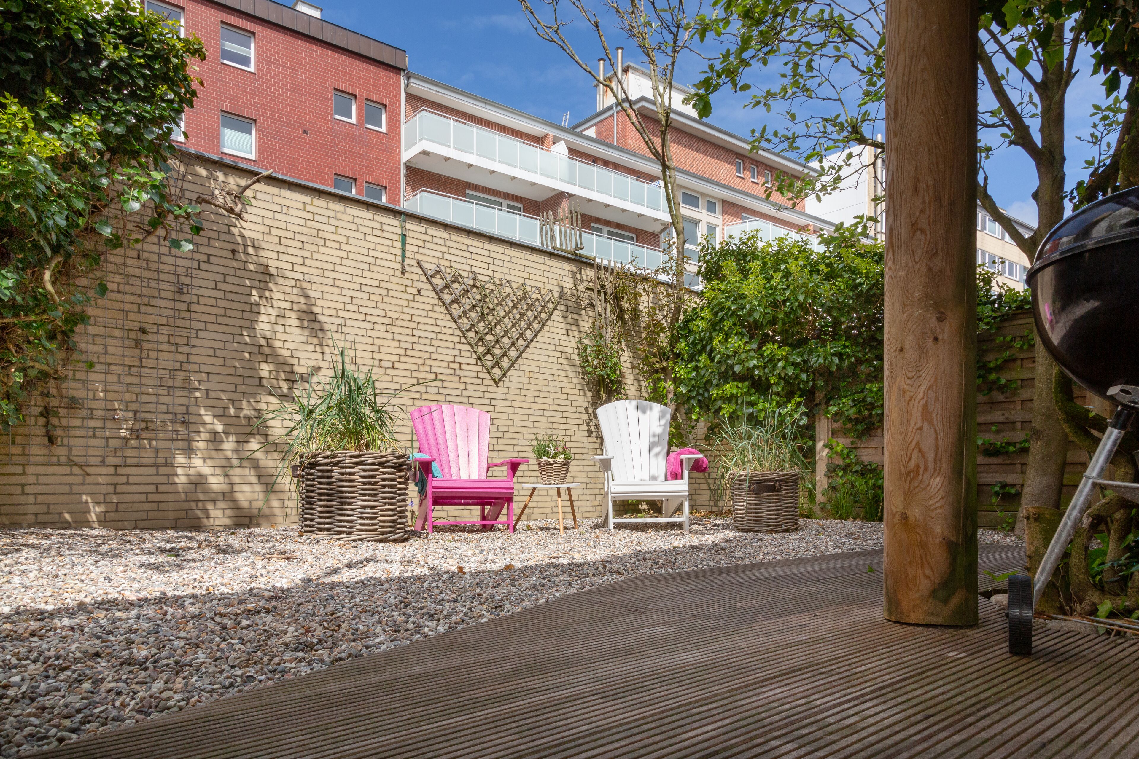 Un patio con piso de madera, dos sillas Adirondack, una parrilla y un muro de ladrillo.