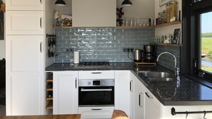 Una cocina moderna con armarios blancos, encimera negra y salpicadero de baldosas grises. Se observan dos lámparas colgantes, una estufa, un horno y un lavaplatos.